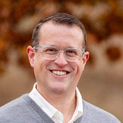 Portrait of James Christopherson, smiling, wearing clear-framed glasses and a grey sweater over a white collar